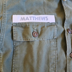 May include: A green utility shirt with a white patch that reads "MATTHEWS" on the left chest pocket.