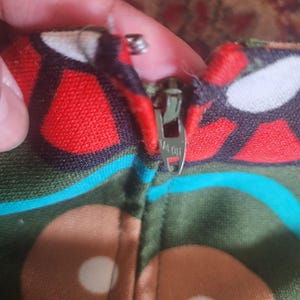 May include: Close-up of a colorful fabric with a zipper. The fabric features a pattern of red, brown, and white shapes on a green background. The zipper is silver and has the word "TALON" printed on it.