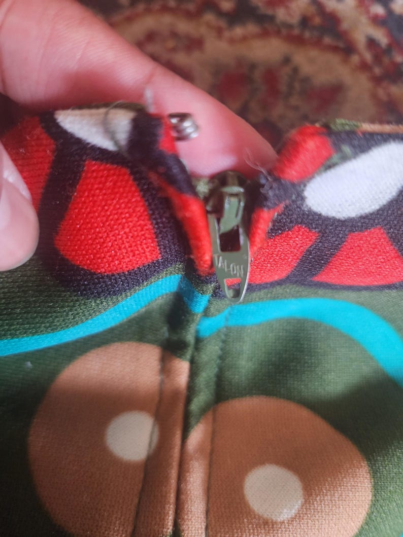 May include: Close-up of a colorful fabric with a zipper. The fabric features a pattern of red, brown, and white shapes on a green background. The zipper is silver and has the word "TALON" printed on it.