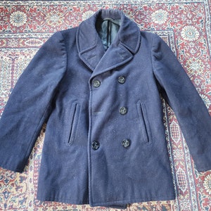May include: A dark blue peacoat with a double-breasted closure and four black buttons. The coat has two flap pockets on the front and a wide collar.