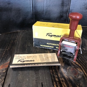 May include: A red and silver automatic numbering machine with a brown handle. The machine is in its original box, which is yellow with black text that reads "Faymus Quality Automatic Numbering Machine". There is also a small instruction booklet that reads "How to operate and care for your Faymus Automatic".