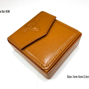 May include: A brown leather case for in-ear headphones. The case has a flap with a stitched design and a logo embossed on the front. The case measures 7cm by 7cm by 2.5cm.