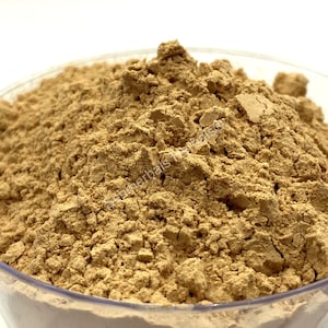 May include: A close-up view of a pile of light brown powder in a clear glass bowl. The powder is finely ground and has a slightly grainy texture.
