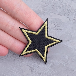 May include: Black star-shaped patch with gold embroidered outline. The patch is made of fabric and has a simple, clean design. The patch is a decorative item, suitable for clothing or accessories.