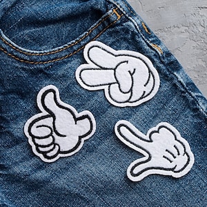 May include: Three white felt hand patches with black outlines sewn onto a blue denim fabric. The patches are in the shapes of a thumbs up, a peace sign, and a pinky finger pointing up.