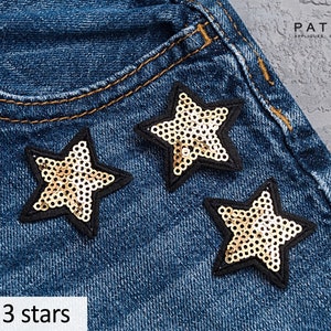 Op de afbeelding: Set van drie gouden, stervormige strijklabels met zwarte randen op een blauwe denim achtergrond. De tekst "Set of 3 stars" staat onderaan de afbeelding.
