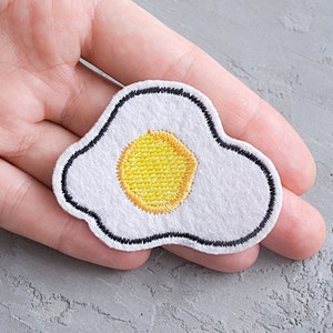 May include: White felt embroidered patch shaped like a fried egg with a yellow yolk.