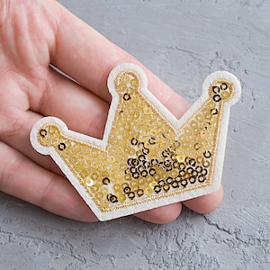 Może przedstawiać: Naszywka w kształcie korony z złotych cekinów z białym filcowym podkładem. Korona ma klasyczny wzór z pięcioma punktami.