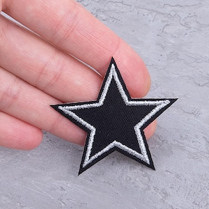 May include: Black star-shaped patch with white embroidered outline. The patch is a five-pointed star with a solid black center. The patch is likely an applique or iron-on for clothing or accessories.