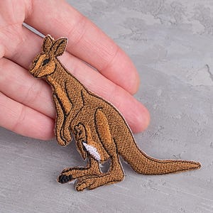 Può includere: Toppa ricamata raffigurante un canguro marrone con marsupio, dettagliata con accenti marrone chiaro e bianchi. La toppa è progettata con un contorno dettagliato ed è probabilmente destinata all'applicazione su abbigliamento o accessori. La toppa è su una superficie grigia.