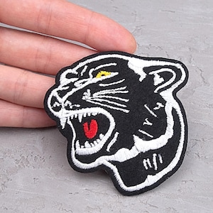 May include: Black and white embroidered patch of a panther's head with a red tongue and yellow eye. The patch is on a black fabric background.