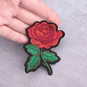 Op de afbeelding: Geborduurde rode rozenpatch met groene bladeren en een zwarte rand. De patch heeft gedetailleerde stiksels, die de bloemblaadjes en bladeren laten zien. De patch is ontworpen voor toepassing op kleding of accessoires.