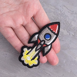Può includere: Un toppa ricamata colorata da stirare che rappresenta un razzo di cartone animato con un design rosso, bianco e blu. Il razzo sta decollando con una fiamma gialla.