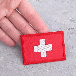 May include: A red embroidered patch with a white Swiss cross. The patch is on a gray surface.