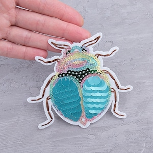 May include: A colorful sequined beetle patch with a white border. The beetle features a rainbow-colored head, a black and white polka-dotted midsection, and turquoise blue wings. The patch is labeled "PATCHFUL" in red text.