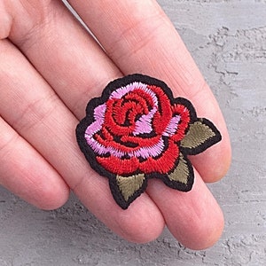 May include: A red and pink embroidered rose patch with green leaves and a black outline.
