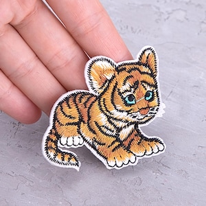 Puede incluir: Parche bordado de un tigre bebé con rayas naranjas y negras, ojos azules y detalles blancos. El parche está en posición agachada, con el nombre de la marca "PATCHFUL" en la parte inferior.
