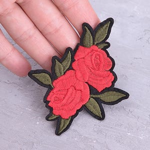 Puede incluir: Parche bordado con dos rosas rojas con hojas verdes y un borde negro. El parche es de Patchful, una marca especializada en apliques y parches. El diseño es adecuado para ropa o accesorios.