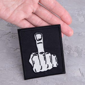 以下が含まれることがあります： グレーの表面に置かれた、白い中指が空中に突き上げられた黒い刺繍パッチ。