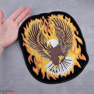 May include: Embroidered patch featuring a majestic eagle with outstretched wings, set against a backdrop of stylized flames. The eagle has brown and white feathers, a yellow beak, and is surrounded by orange and yellow flames on a black background.
