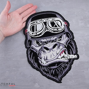 Puede incluir: Parche bordado con un gorila que lleva un casco y gafas negros, con un cigarro. El gorila tiene una expresión severa, con costuras detalladas en tonos grises, negros y blancos. El parche está sujeto por una mano.