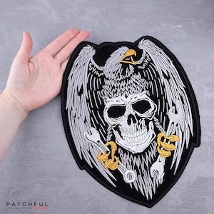 Puede incluir: Parche bordado negro con un cráneo con un águila encima y llaves inglesas a cada lado. El águila es gris y blanca, el cráneo es blanco y las llaves son plateadas y doradas. El parche mide aproximadamente 20 cm de ancho.