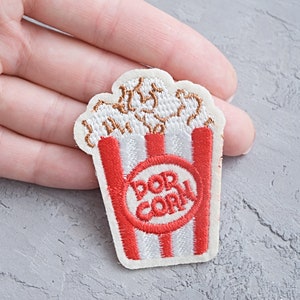 Pode incluir: Etiqueta branca bordada para passar a ferro de um balde de pipocas com riscas vermelhas e brancas com a palavra "Popcorn" num círculo.