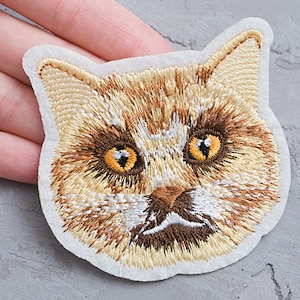 May include: Embroidered iron-on patch of a cat's face with brown and white fur and yellow eyes.