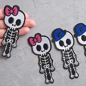 Può includere: Quattro toppe ricamate da stirare con figure di scheletri con diversi accessori. Le toppe sono bianche con contorni neri e dettagli rosa e blu.