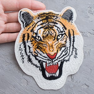 Op de afbeelding: Een geborduurde strijk-patch met het gezicht van een tijger in oranje, zwart en wit. De tijger heeft een woeste uitdrukking met een wijd open mond en scherpe tanden.