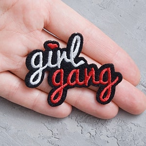 Può includere: Un cerotto in tessuto nero con ricamo bianco e rosso che recita "girl gang".