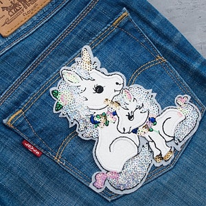 Può includere: Un paio di toppe di unicorno bianche con paillettes iridescenti, una più grande dell'altra, cucite su una tasca di jeans blu. L'unicorno più grande ha un motivo floreale intorno al collo.