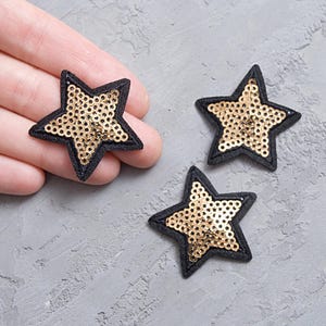 Può includere: Tre toppe a forma di stella. Ogni toppa ha un bordo nero ed è riempita di paillettes dorate. Le toppe sono su una superficie grigia. Una toppa è tenuta in mano.