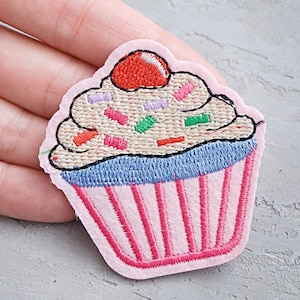 Puede incluir: Un parche bordado de un cupcake rosa con glaseado blanco, una cereza roja y espolvoreado de colores.