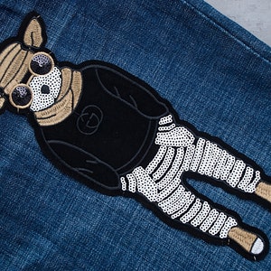 May include: Embroidered patch featuring a dog-like character wearing sunglasses and a black outfit with a skeleton-style design on the legs. The patch is on a blue denim background.