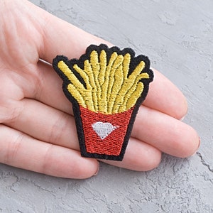 以下が含まれることがあります： 赤い白の紙コップに黄色いフライドポテトが入った刺繍のアイロン接着パッチ
