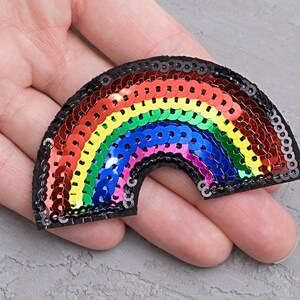 以下が含まれることがあります： 虹色のスパンコールパッチで、アーチ型をしています。パッチは、黒い布地に縫い付けられた小さな虹色のスパンコールでできています。