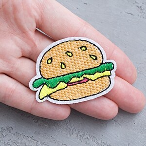 Pode incluir: Etiqueta bordada para passar a ferro de um desenho animado de um hambúrguer com alface, queijo e pão de sementes de sésamo. A etiqueta é branca com bordado amarelo, verde e castanho.