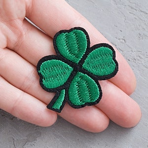 Può includere: Una toppa ricamata a forma di trifoglio a quattro foglie di colore verde con cuciture nere.