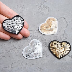 Può includere: Quattro toppe a forma di cuore con paillettes. Due toppe sono nere con paillettes argentate, una è bianca con paillettes argentate e una è bianca con paillettes dorate.