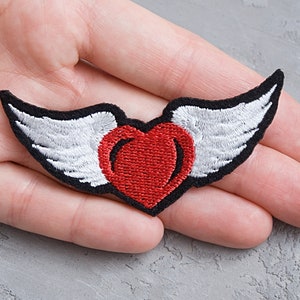 Peut inclure: Patch brodé représentant un cœur rouge avec des ailes blanches. Le cœur est centré et les ailes sont déployées de chaque côté.