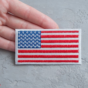 May include: A white embroidered patch with the American flag design. The flag has 50 white stars on a blue background and 13 red and white stripes.