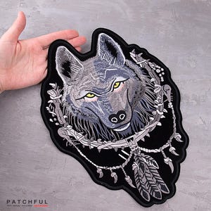 Peut inclure: Écusson brodé de loup avec des détails gris et noirs, avec des yeux jaunes et une bordure décorative. L'écusson a un fond noir et mesure environ 20 cm de large.
