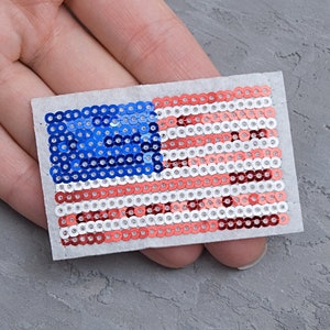 May include: A small American flag patch made of red, white and blue sequins. The flag is on a white background.