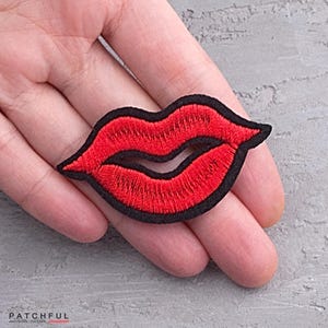 以下が含まれることがあります： 黒い輪郭のある赤い刺繍の唇のパッチ。パッチの幅は約5cm、高さは約2.5cmです。