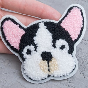 May include: A white, black and pink felt patch of a French bulldog's face. The patch is designed to be ironed onto fabric.