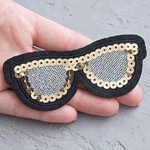 Puede incluir: Un parche de tela negro con forma de gafas de sol con lentejuelas doradas alrededor de las lentes.