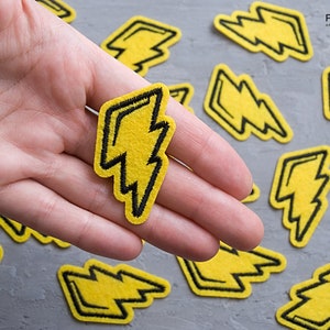 May include: Yellow felt lightning bolt patches with black stitching. The patches are approximately 5 cm tall and are arranged on a grey surface.