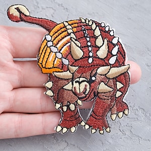 May include: Embroidered patch of an Ankylosaurus dinosaur. The patch features shades of brown, orange, and yellow, with white accents. The dinosaur is depicted with spiky armor and a club tail, perfect for adding to clothing or accessories.