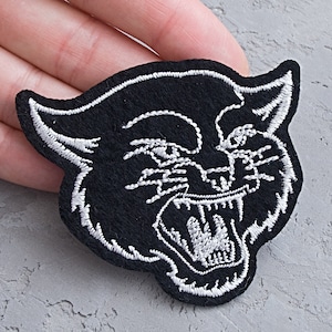May include: Black embroidered iron-on patch of a snarling cat's face with white stitching.